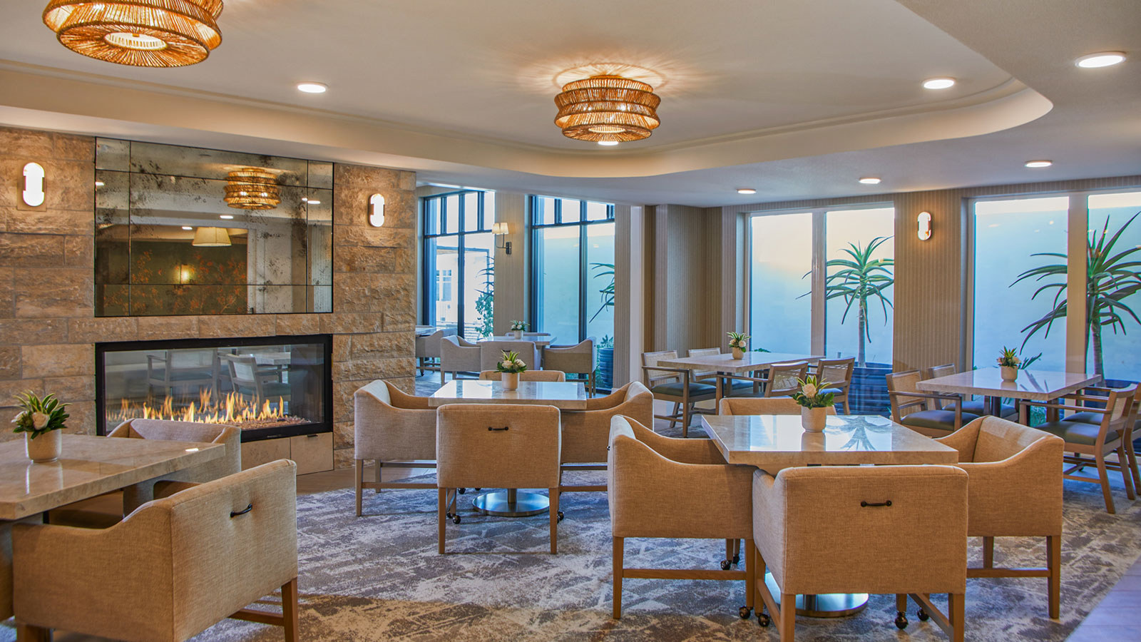 This is a photo of The Seville's assisted living dining room. The room is filled with chairs, features a fireplace, and has floor-to-ceiling windows.